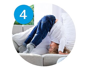 A child is bending forward on a couch, with their head down, wearing a white shirt and blue jeans.