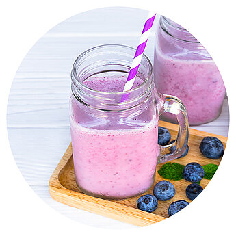 A transparent glass jar filled with a pinkish smoothie, accompanied by a purple and white striped straw, sits on a wooden tray alongside scattered blueberries and a mint leaf.