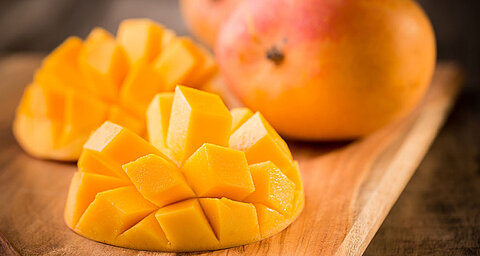 A mango, cut into precise cubes, sits on a wooden cutting board, with a whole mango in the background.