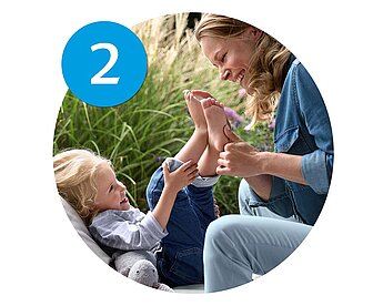 A woman and a child are seated outdoors, with the woman holding the child's foot and looking at it.