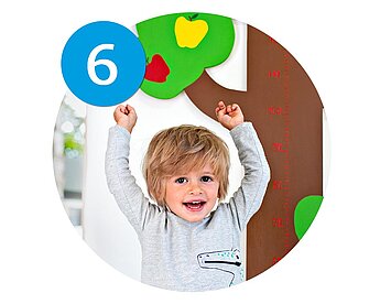 A child stands in front of a height measurement chart, raising their arms in excitement.