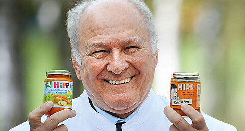 An elderly man with a white shirt holds two jars of baby food, one with a label that reads "HIPP Karotten" and the other with "HIPP Frukt-Karotten-Gemüse-Kartoffeln."
