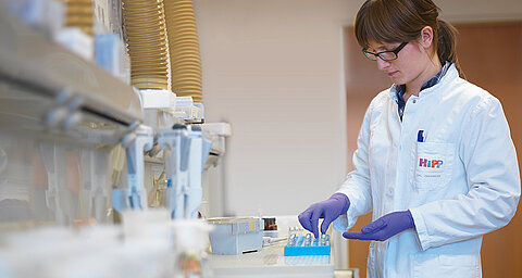 A person in a white lab coat is working with a blue tray containing small items, surrounded by laboratory equipment.