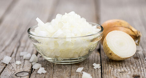 A transparent bowl filled with finely chopped onions sits on a wooden surface, with a whole onion and its halved counterpart nearby.