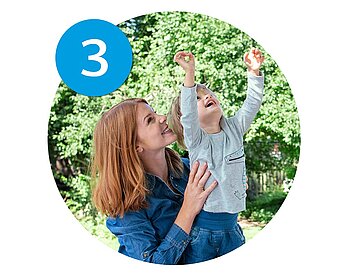 A woman in a blue shirt holds a child in a gray shirt, both looking up with their arms raised, against a backdrop of green trees.
