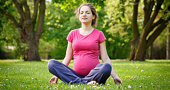 A woman in a pink shirt and blue pants sits cross-legged on a grassy field with flowers, surrounded by trees.