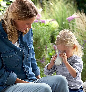 A woman and a young girl sit outdoors, with the girl drinking from a glass.