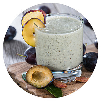 A glass of creamy beverage is garnished with a plum slice, surrounded by plums and almonds on a wooden surface.