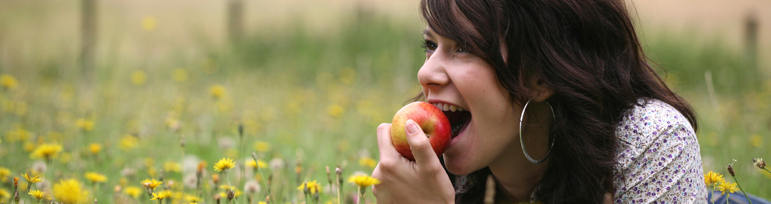 A woman with long dark hair is biting into a red apple while lying in a field of green grass and yellow flowers.