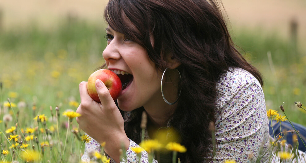 A woman with dark hair and a floral shirt is biting into a red apple while lying on grass surrounded by yellow flowers.