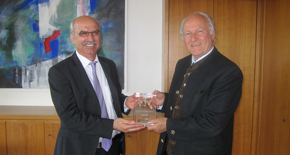 Two men in suits stand in a room with a painting and wooden cabinets. One man presents a clear award with text to the other.