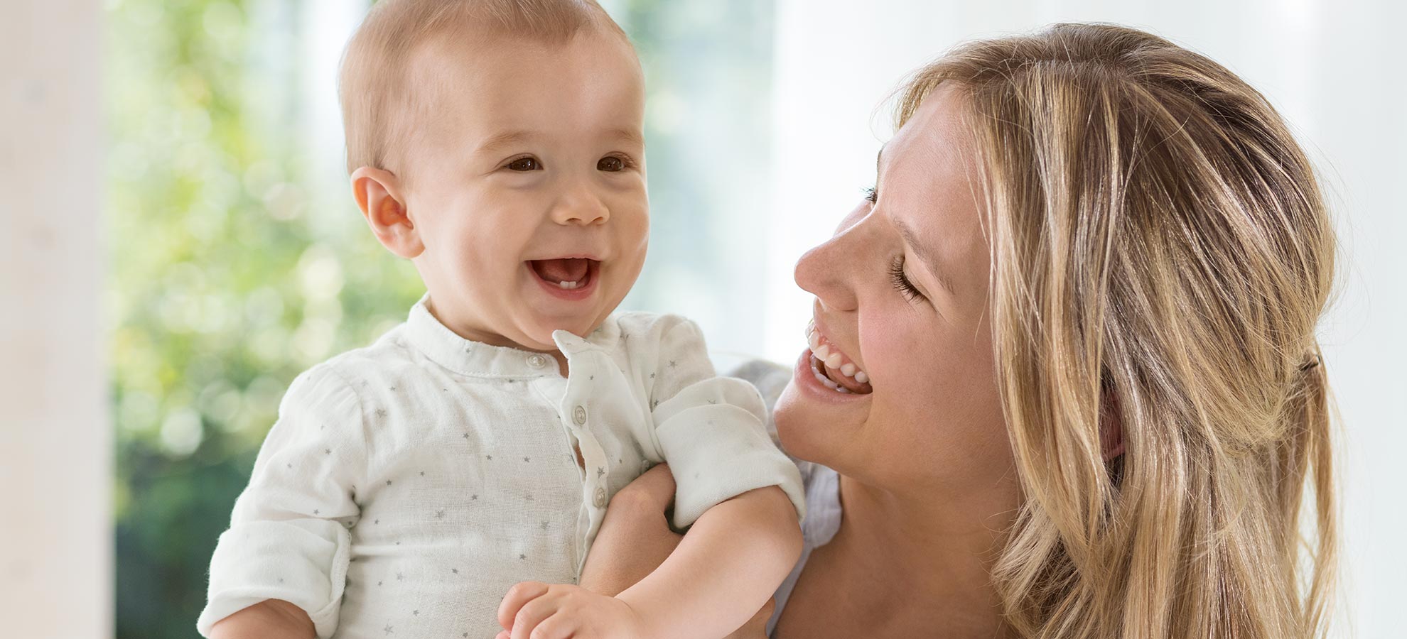 A woman is holding a baby, both looking at each other with smiles.