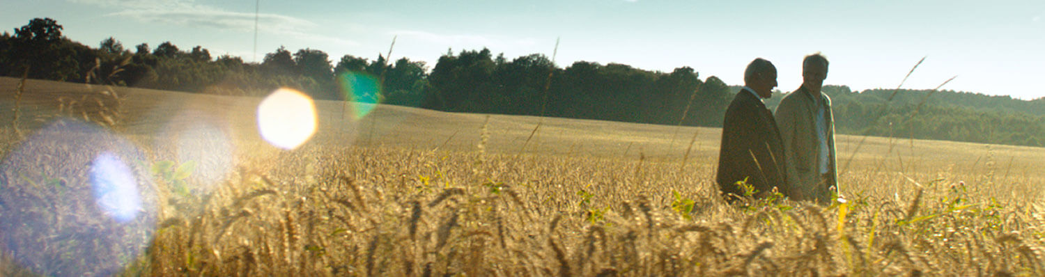 Two individuals stand amidst a field of tall grass, with a forested area in the background under a clear sky.