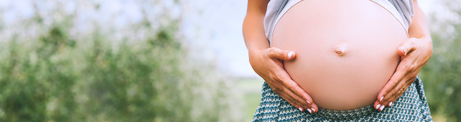 A pregnant woman stands outdoors, her hands gently cradling her belly.