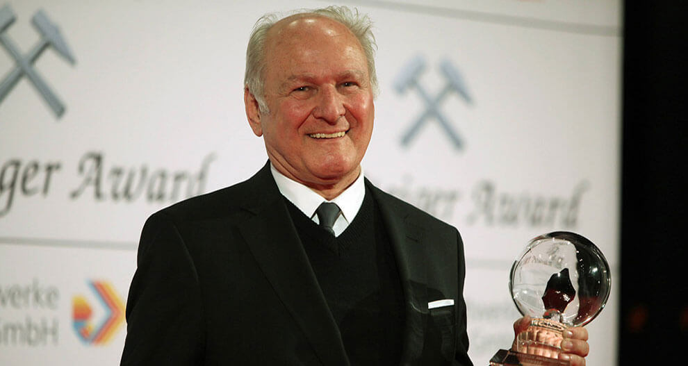 An elderly man in a black suit stands on a stage, holding a transparent award with a silhouette of a person, against a backdrop that includes the words "Schweizer Award" and a logo.