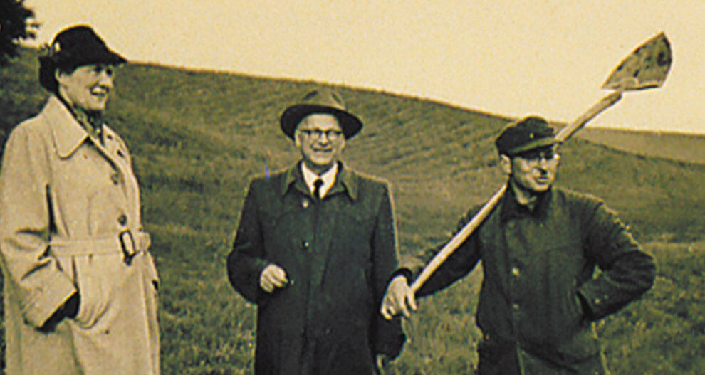 Three individuals stand in an open field; one holds a shovel.