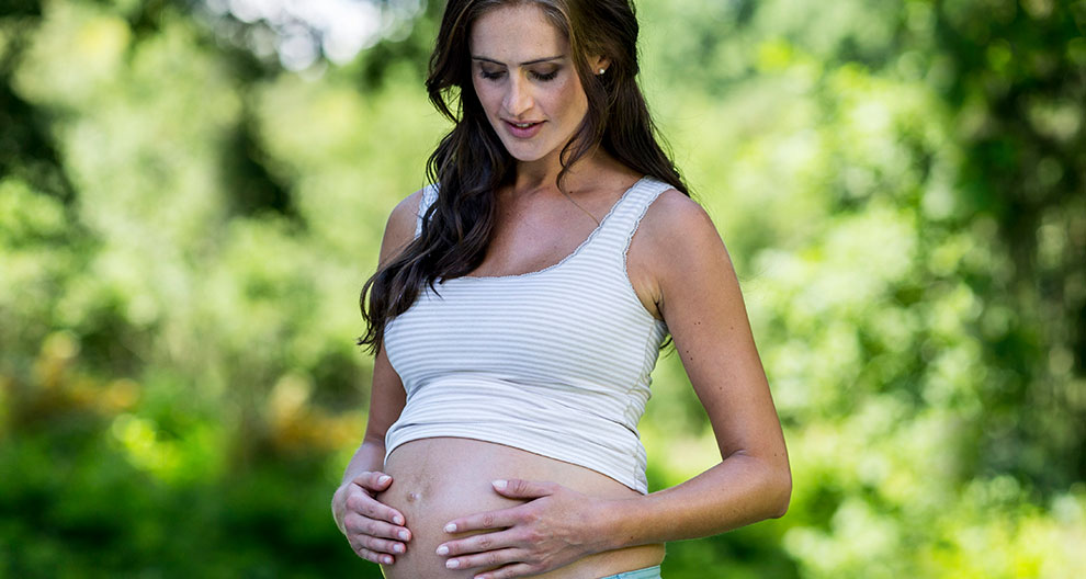 A pregnant woman in a white tank top and blue jeans stands outdoors, holding her belly gently.