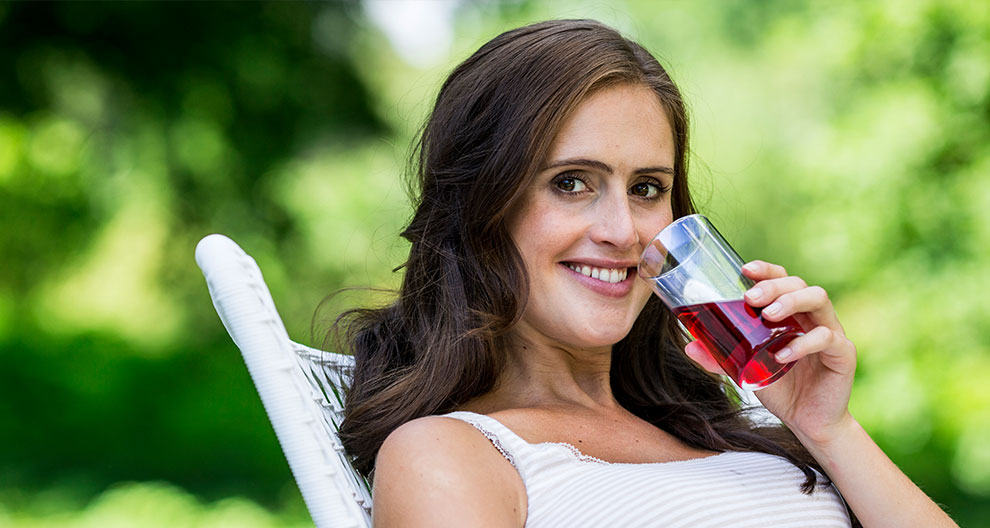 A woman with long brown hair is sitting on a white chair, holding a glass of red liquid, with a green outdoor background.