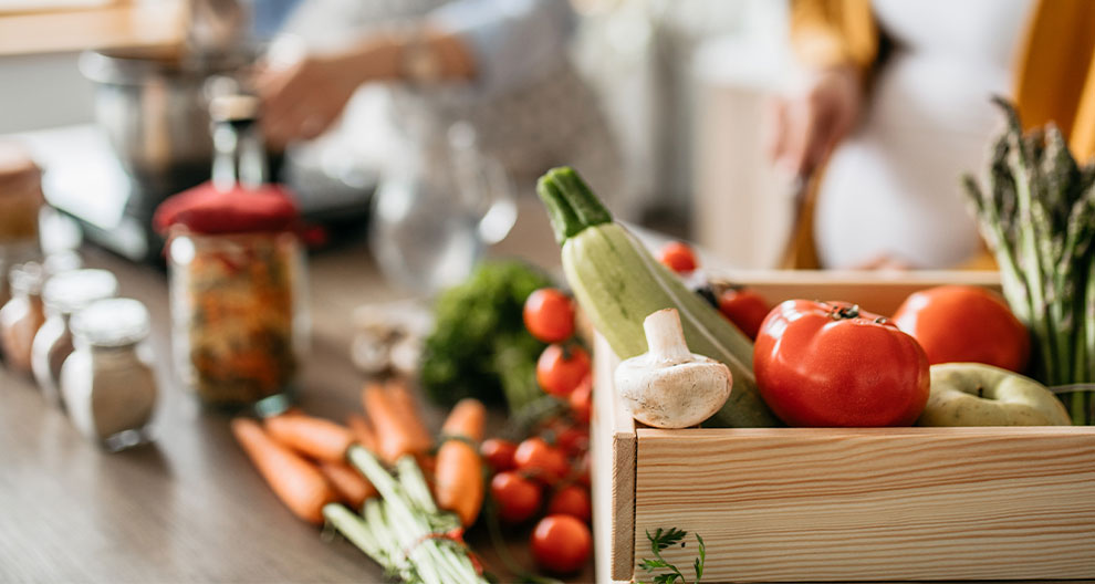 A wooden box filled with fresh vegetables is placed on a table, with a person in the background preparing food.