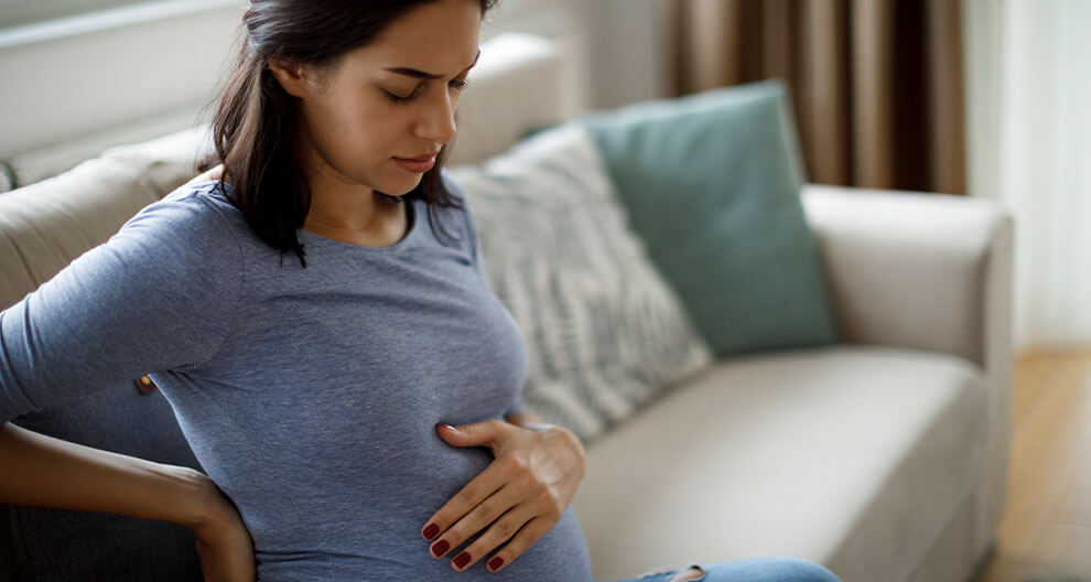 A pregnant woman in a blue shirt sits on a couch, her hand resting on her belly.