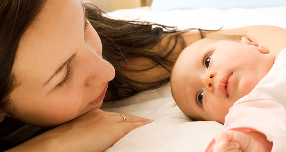 A woman and a baby are lying on a bed, looking at each other.