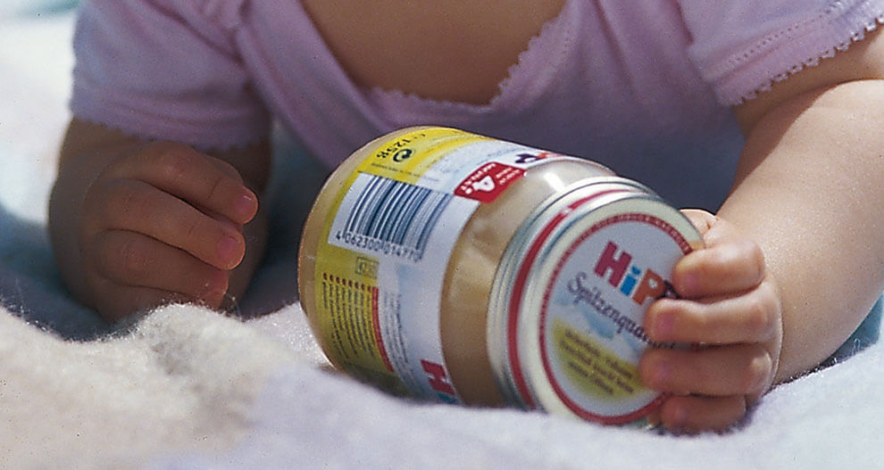 A baby's hand grasps a jar with a barcode and label, set against a soft, white background.