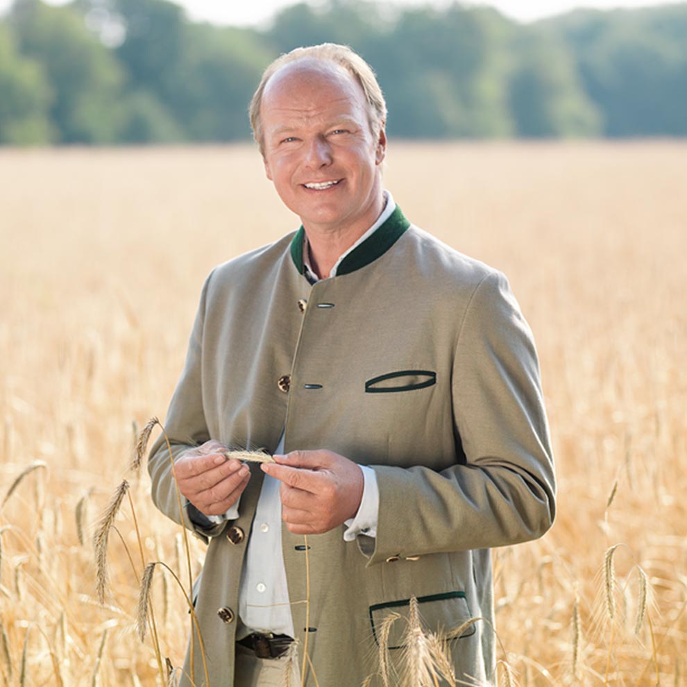 A man in a green jacket stands in a wheat field, holding a wheat ear.