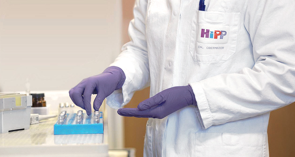 A person in a white lab coat with the logo "HiPP" and the name "CH. OBERMAIER" is handling a blue tray with multiple small containers.