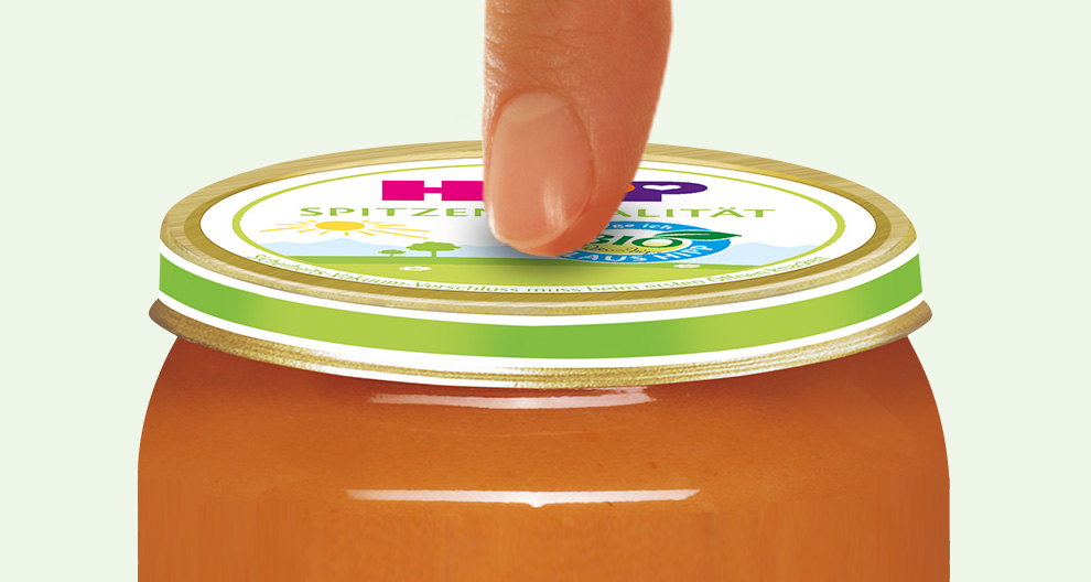 A finger is about to open a jar with a green lid, containing a brownish-orange substance, with a sticker on the lid that reads "BIO" and other text.