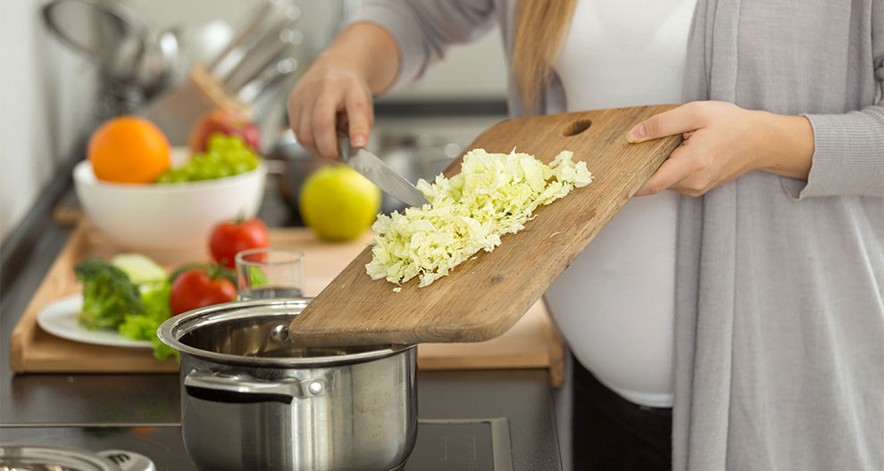 A person is chopping cabbage on a cutting board, with a pot nearby, and various fruits and vegetables in the background.