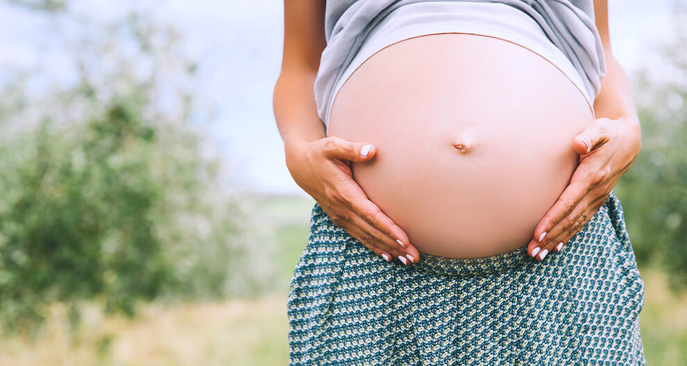 A pregnant woman stands outdoors, her hands gently cradling her belly.