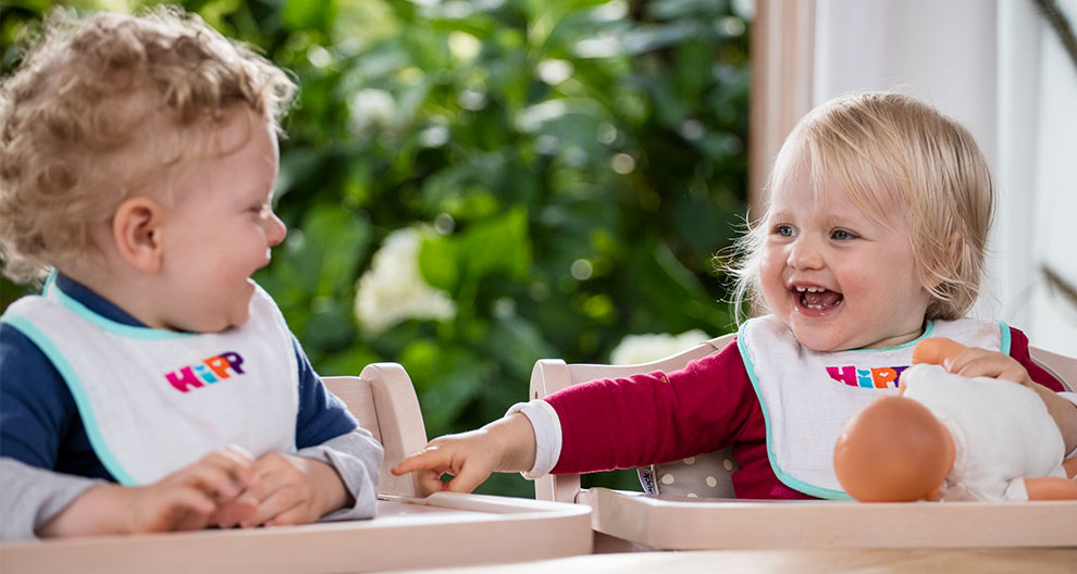 Two toddlers, one in a blue shirt and the other in a red shirt, sit across from each other, both wearing bibs with the word "HIP" on them, and the toddler in red is holding a doll.