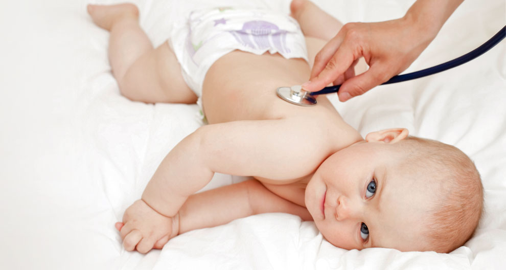 A baby lies on a white sheet while a hand places a stethoscope on the baby's back.