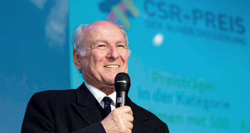 An elderly man with a bald head and a white beard is holding a microphone, standing in front of a blue background with white text.